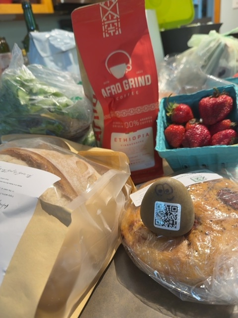 A small toy figure would be resting on a white countertop or table surface surrounded by fresh groceries and packaged goods in what appears to be an indoor kitchen or market setting during daytime. The figure would be positioned on or near a loaf of bread with a QR code sticker, surrounded by fresh strawberries in a blue container, leafy greens in plastic bags, a red coffee package, and other food items, with bright overhead lighting creating clear visibility. No people are visible in the frame, which captures an organized arrangement of produce and packaged foods at close range.