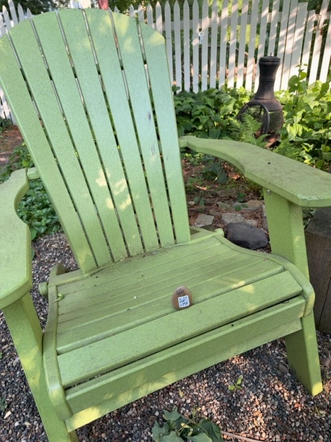 A small toy figure appears to rest on the seat of a bright green Adirondack chair positioned in a gravel garden area. The chair sits on a bed of small stones with garden beds and white picket fencing visible in the background, suggesting a residential yard during daytime with dappled sunlight. The perspective is at ground level, with the chair's slats and armrests dominating the view, and various outdoor objects including plants, garden equipment, and landscaping elements visible in the soft-focused background.