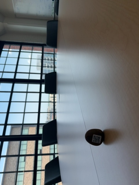 A small dark object rests on a white ceiling or overhead surface, viewed from below in bright daylight streaming through a large window with a black grid frame. The window overlooks an urban or industrial area with multi-story buildings visible in the distance, and the scene is photographed indoors from a tilted angle that emphasizes the height and expanse of the plain white space above.
