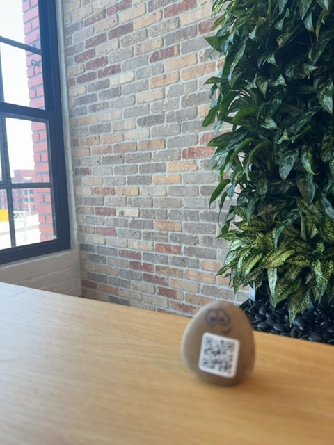A small painted rock with a QR code sits on a light wooden table surface in what appears to be an indoor office or commercial space. Behind it, a brick wall with warm reddish and tan tones stretches upward, with a black-framed window on the left and a lush green living wall plant installation on the right. The natural daylight streaming through the window suggests daytime hours, and the setting feels modern and carefully designed, with the painted rock being the only toy-like object visible in the frame.