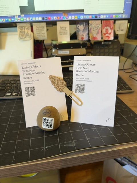 A small painted rock with a QR code rests on a cutting mat in the foreground, positioned between two printed documents and a woven plant hanger with a golden tone. Behind the rock, a desk workspace is visible with a computer monitor displaying a taskbar, various wooden boxes or blocks arranged on a shelf above the monitor, and computer peripherals including a keyboard and mouse. The setting appears to be indoors during daytime with artificial lighting, creating a workspace environment where someone has arranged these objects for documentation or display purposes.