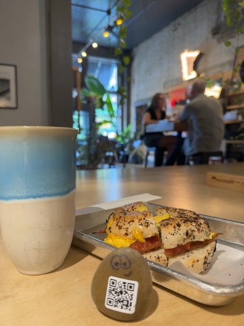 A small painted stone figure with a simple face rests on a wooden counter in the foreground, positioned between a two-toned ceramic mug and a seeded bagel sandwich on parchment paper. The setting is a bright, modern café interior with warm string lights strung overhead, green plant vines, and a concrete-textured back wall lit by neon signage, suggesting early evening hours. In the soft-focus background, several people sit at tables and the counter, engaged in conversation, while the overall space feels open and contemporary with natural wood surfaces and industrial-style finishes.