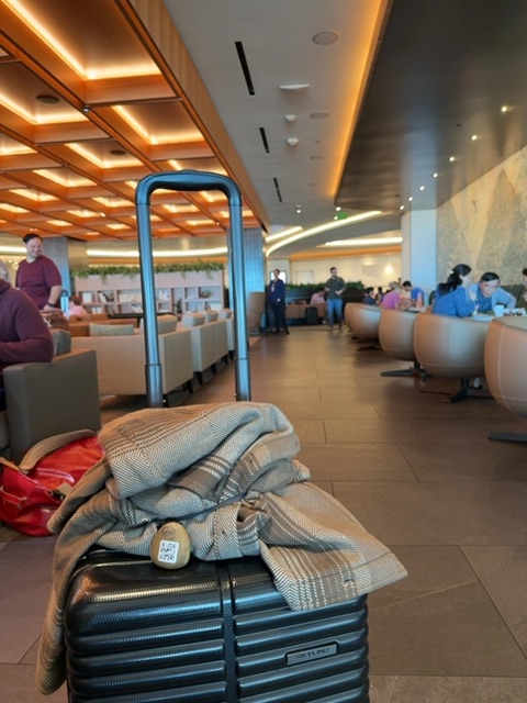 A small painted rock with two round eyes and a QR code is resting on top of a black ridged suitcase in the foreground, among folded textiles and a red item. The suitcase sits on a tan-colored floor in a large interior space with modern architectural features including wooden ceiling panels with warm orange lighting, blue metal railings, and cylindrical seating pods distributed throughout. Multiple people are scattered throughout the space in the background at various distances, some standing and some seated, with a doorway or opening visible on the left side. The overall setting has warm and cool lighting tones, appears to be indoors in a contemporary commercial or transit space, and conveys a sense of scale suggesting an expansive, enclosed room.