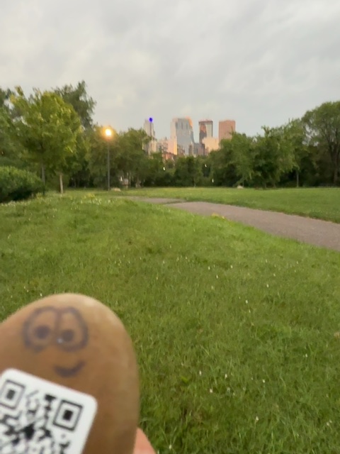 A small painted brown figure with a simple smiling face is held up in someone's hand in the foreground of this photo, positioned to look out toward a park setting. Behind it stretches a wide open green lawn with a curved pathway, tall trees lining both sides, a street lamp glowing on the left, and a distant cityscape of tall buildings visible on the horizon under an overcast sky. The lighting suggests early evening or dusk, with warm artificial light beginning to activate in the urban background.