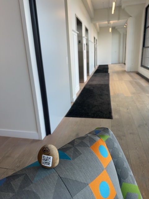 A small wooden toy figure with a QR code face rests on a colorful patterned cushion or fabric object in the foreground, positioned at floor level in a modern hallway. The hallway stretches behind it with white walls, closed doors on the left side, fluorescent ceiling lights, and a dark runner rug down the center, creating a perspective that emphasizes the toy's small scale relative to the architectural space. The lighting is bright and neutral, typical of daytime in an indoor office or institutional setting, with no people visible in the frame.