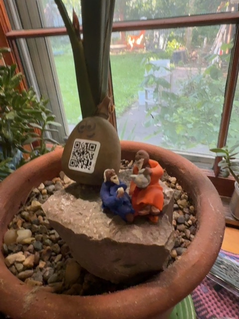 A small toy figure in blue and orange clothing stands on a painted rock within a terracotta pot filled with small stones and sand, positioned on a windowsill. Behind the pot, large windows frame a garden with green grass and planted areas visible in natural daylight. A hand belonging to someone outside the frame holds or steadies the pot, and various indoor plants and objects are visible around the windowsill setting.