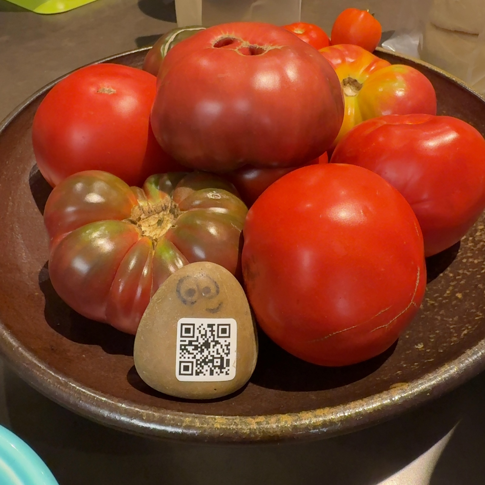 A painted river rock with two round eyes and a QR code sticker rests in the foreground of a brown ceramic bowl, positioned among several tomatoes of varying sizes and colors. The bowl sits on what appears to be a neutral-toned surface, with blurred background elements including light-colored vertical structures and a green object in the upper left. The tomatoes are predominantly bright red, with some darker reddish-brown varieties visible, and their varied sizes create an uneven arrangement around the small painted rock. The lighting is bright and natural, casting clear shadows and highlighting the glossy surfaces of the tomatoes and the ceramic bowl.