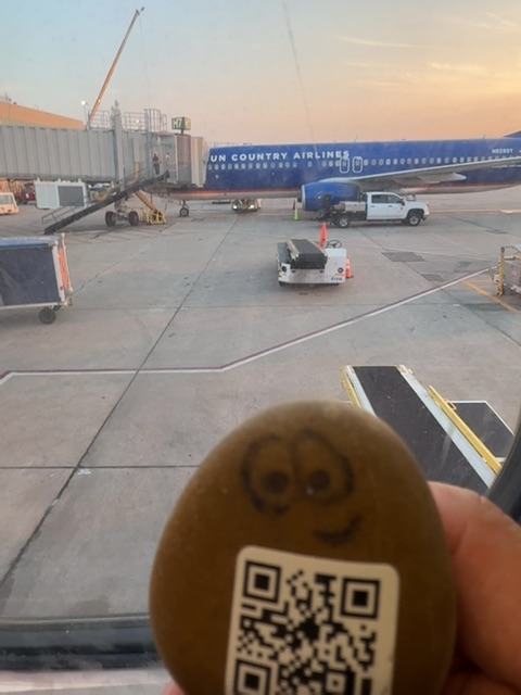 A small painted or toy figure with a QR code on its base is held in someone's hand in the foreground, with an airport tarmac stretching behind it during golden hour at dusk. The scene shows a blue commercial aircraft, ground service vehicles, boarding equipment, and a terminal building illuminated by warm sunset light. The figure's vantage point from a person's hand creates a dramatic scale perspective over the expansive, mostly empty concrete apron.