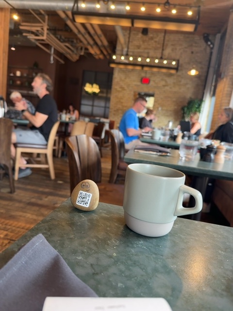 A small painted rock with a QR code rests on a dark green table surface next to a white ceramic mug in what appears to be a modern restaurant or café interior. The space features warm overhead lighting, wooden chairs, brick walls, and several people seated at tables in the background engaged in conversation. The vantage point is from table level, looking across the mug toward the softly lit dining area with its contemporary décor and furnishings.