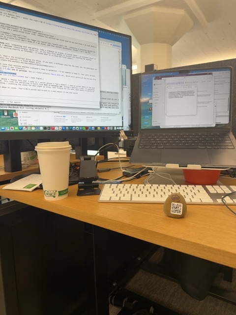 A small toy figure would be resting on a wooden desk surface, positioned near a coffee cup and keyboard in a workspace with dual monitors displaying documents and software interfaces. The space is brightly lit by overhead ceiling lights, creating a daytime indoor office setting enclosed by beige walls and dark furniture, with a laptop visible to the right and various desk items surrounding the figure's vantage point. No small toy figures or painted rocks are visible in the frame itself.