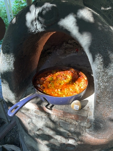 A small toy figure rests on the rim of a blue cooking pot containing a bubbling orange stew or bean dish, positioned inside a large concrete or stone oven with a dark interior and rusty metal opening. The setting appears to be outdoors in daylight with dappled shadows falling across the oven's surface, and a person's hands are visible at the edges of the frame tending to the cooking. The scene conveys a rustic outdoor cooking setup with strong contrast between the bright sunlit areas and the dark oven opening.