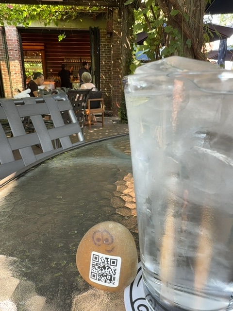 A painted river rock with two round eyes and a QR code label rests on the ground in the foreground of this image. Behind it stands a tall translucent cylindrical object, and further back is a curved gray latticed barrier or fence. The scene is set in a paved courtyard or alley area with brick buildings on the left side, where a wooden door and climbing vines are visible, and several people can be seen near the doorway in the middle distance. Dappled sunlight filters through overhead vegetation, creating a mix of light and shadow across the textured ground and structures, suggesting a sunny afternoon in an enclosed urban courtyard space.