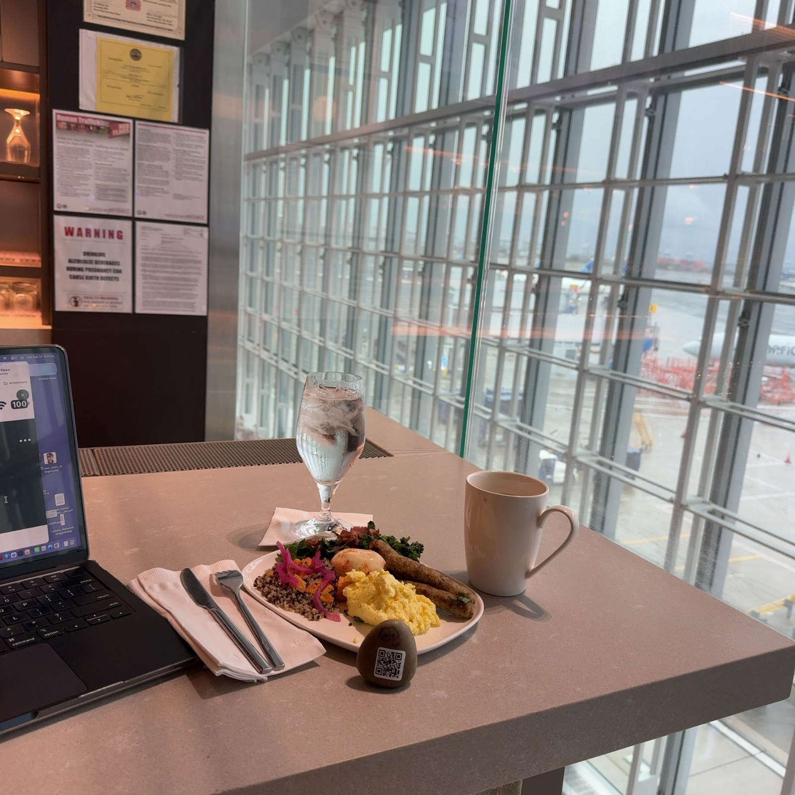 A small painted rock with two round eyes and a QR code marking its face sits on a beige table in the foreground, positioned near a plate of food and dining utensils. The table is situated before a large gridded window that opens onto an expansive view of a waterfront with shipping containers and industrial structures visible in the distance. To the left of the rock sits a laptop computer, a white mug, a glass with ice and liquid, and a QR code reader device on the table surface. The scene is brightly lit by natural daylight streaming through the extensive window panels, which reveal an overcast sky and industrial port landscape beyond, creating a sense of considerable height and openness.