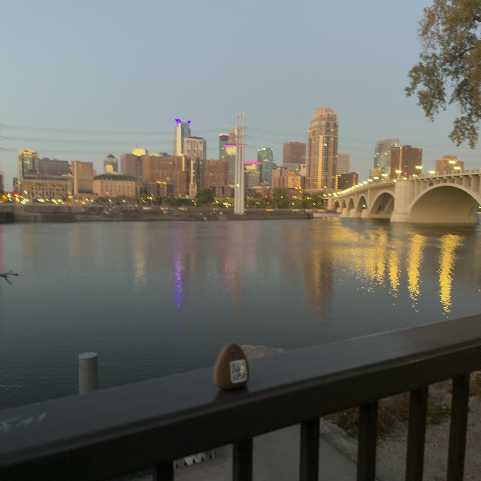 A small brown, rounded river rock with a square QR-code sticker on its front sits on top of a dark metal railing in the lower center of the frame; I can't clearly see painted eyes, but the sticker matches the hint. Beyond the railing, a wide stretch of calm water spreads out, with a short post at its edge on the left and a paved path and gravel visible below the railing on the right. On the far shore, a cluster of mid-rise and high-rise buildings stands behind a low embankment, with a tall pair of white vertical pylons in the middle and a long, pale, multi-arched bridge with a row of lights extending in from the right. No people are clearly visible. The sky is pale blue-gray, the building windows and bridge lights glow yellow, pink, and purple and reflect in streaks on the water, and a tree branch with muted leaves hangs in the upper right, giving a dusk feel and an open, spacious scale, with the background slightly blurred.
