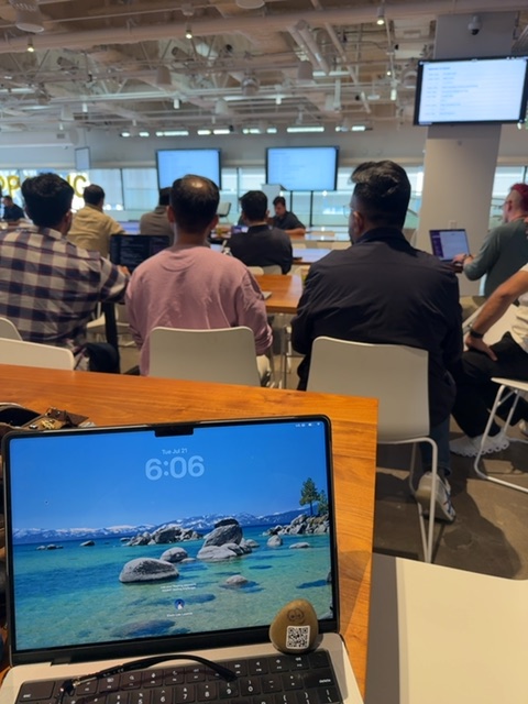 A small toy figure would be resting on a wooden table in the foreground of a bright, modern office or workspace with high ceilings and industrial-style lighting. From this vantage point, the figure would view a laptop screen displaying a scenic landscape, while in the background multiple people in casual clothing stand and move about the open floor plan, some gathered near windows and displays on the far wall. The warm wood of the table contrasts with white chairs and the cool blue tones of the laptop screen and distant windows, suggesting daytime in a professional collaborative environment.