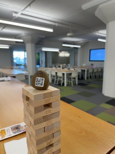 A wooden block with a QR code painted on its spherical top rests atop a stacked wooden tower on a light wood table in a bright, modern office space. The scene is illuminated by fluorescent ceiling panels and natural light from windows, with the vantage point at table height looking across an open floor plan featuring green carpet sections, gray columns, and meeting areas with screens visible in the background. No people are visible in close proximity, though the space appears designed for collaborative work with multiple functional zones receding into the distance.