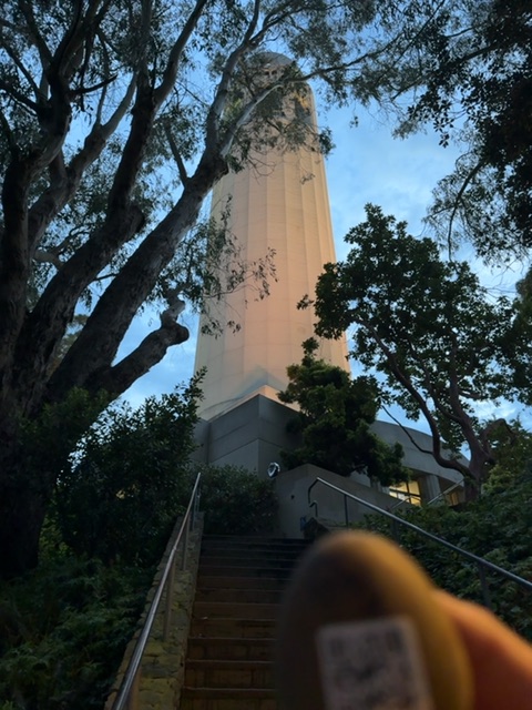 A small tan toy figure with a QR-code face is held close to the camera at the bottom of the frame, slightly out of focus. Beyond it, a long outdoor staircase climbs through dense green vegetation toward a tall pale concrete tower and a dark building at its base. Large trees frame the tower against a blue sky in soft daylight. No people are visible.