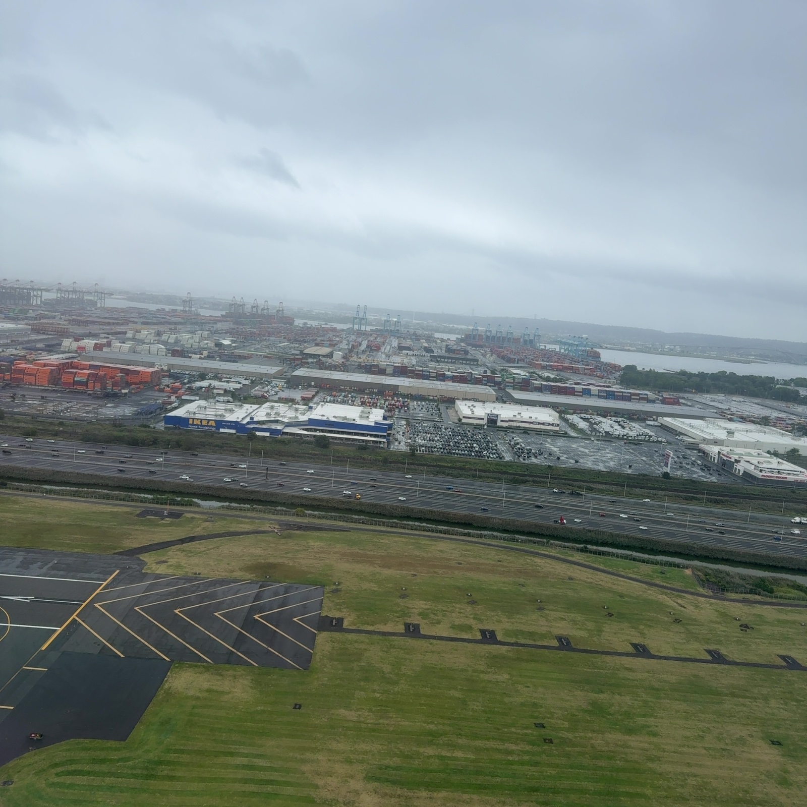 No figure matching the hint—a palm-sized painted rock with eyes and a QR code—is clearly visible in this photograph. The image shows an aerial view taken from an aircraft, with a runway and taxiway marked with yellow chevrons visible in the immediate foreground on dark asphalt, and grassy areas with ground-level structures alongside. Beyond a multi-lane highway with vehicles and a green barrier, industrial port facilities dominate the middle distance, including large shipping containers stacked in red, multiple white warehouse structures, and numerous cargo-handling cranes silhouetted against the overcast sky. Water is visible in the far background where the port meets a body of water, with forested land beyond. The light is diffuse and gray from heavy cloud cover, creating a muted color palette of greens, grays, and industrial colors, with the overall sense of a large, open, industrial landscape viewed from significant height.