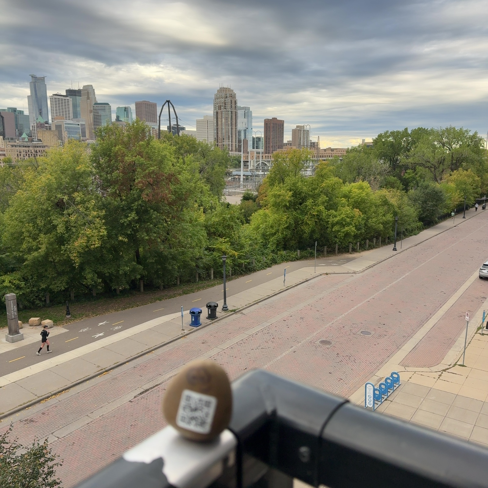A small painted rock with eye markings and a qr code rests on a black metal railing in the foreground, close to the camera and out of focus. Below and beyond it runs a wide brick street with a sidewalk and a bike path, trees lining the path on the left side and green foliage filling the middle distance. Beyond the trees, a sharply focused city skyline rises with multiple tall buildings of varying heights, including a distinctive peaked structure and an arched gateway or monument, set against an overcast sky. A single person is visible in the distance on the lower left, on the path near the trees, and the overall scene conveys an open, elevated viewpoint overlooking an urban park area in daytime under cloudy conditions.