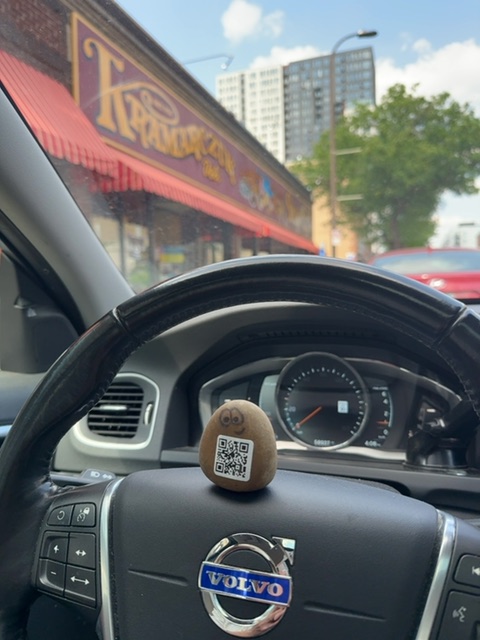 A small object with a QR code printed on it rests on the steering wheel of a Volvo car in daylight, positioned to face the windshield. Through the windshield, a street scene is visible showing a storefront with red and gold signage, apartment buildings, trees, and sky with scattered clouds. The car's interior features a black steering wheel, dashboard with gauges, and air vents, all in sharp focus in the foreground while the exterior urban landscape appears slightly softer behind the glass.