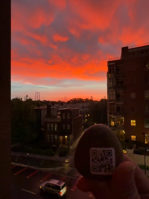 A small toy figure or painted object with a QR code pattern is held in someone's hand against an urban rooftop view during sunset or dusk. The sky displays vivid orange and red clouds while buildings with lit windows frame the scene below, and distant amusement park rides are visible on the horizon. The figure's vantage point is elevated several stories above street level, looking out over a cityscape during the golden hour.