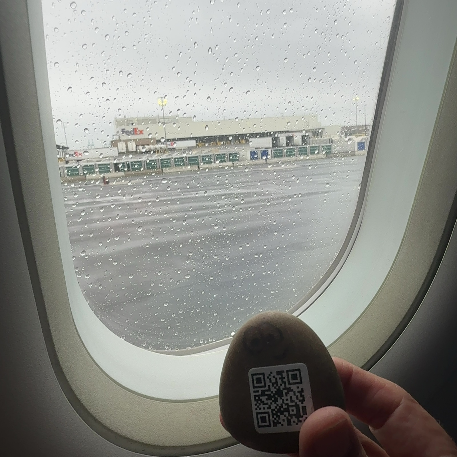 A painted river rock with two round eyes and a QR code is held in someone's hand in the foreground of the frame. Through an airplane window behind it, a wet tarmac surface is visible, covered with water droplets, stretching away toward airport buildings and structures in the distance. A FedEx facility and green boarding bridges or cargo structures are identifiable among the buildings lining the far side of the tarmac. The sky is overcast and gray, and the window itself is heavily beaded with condensation and rain, creating a blurred, wet texture across most of the view.