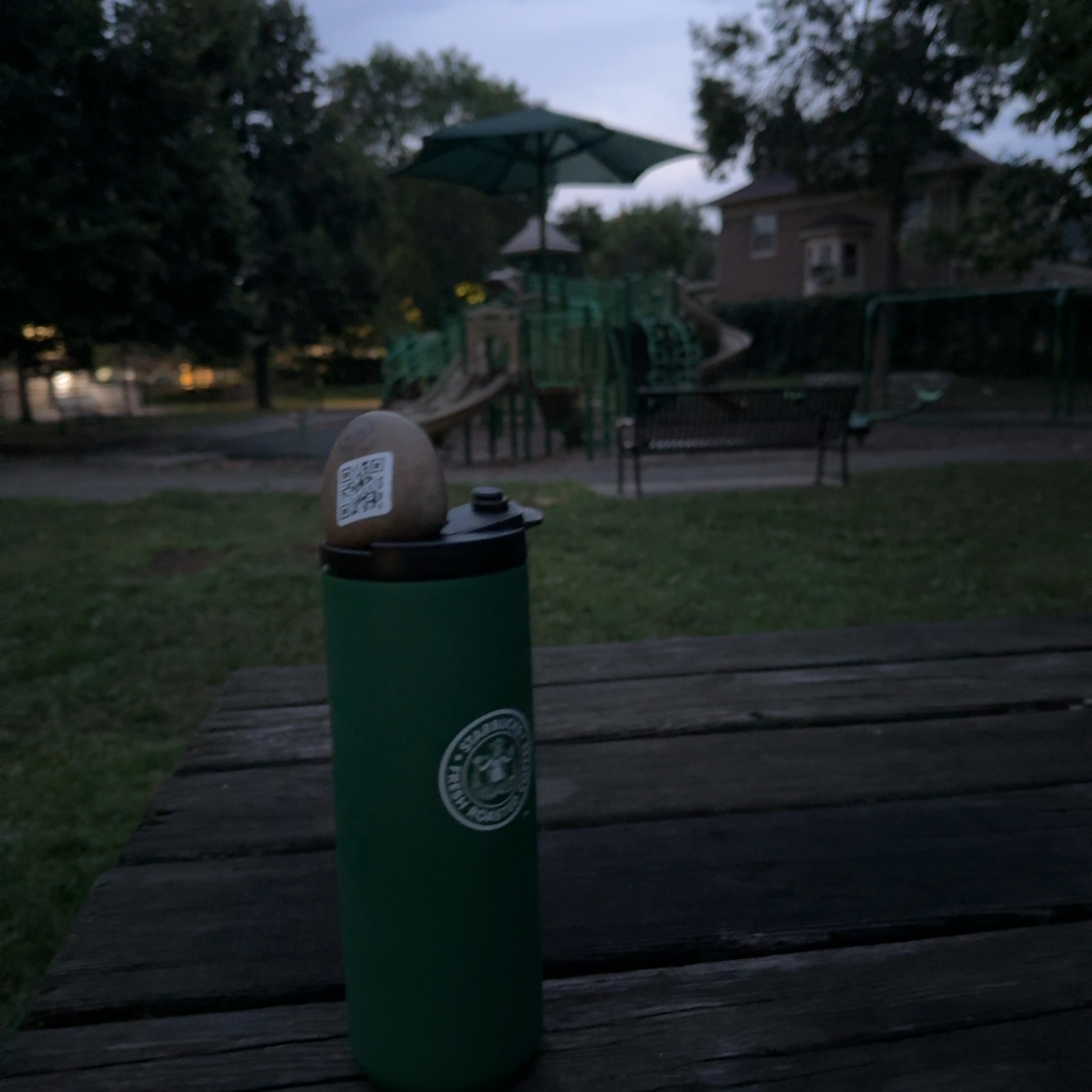 A small painted rock with two round eyes and a QR code rests on top of a green cylindrical post in the immediate foreground, positioned on what appears to be a wooden bench or platform. Behind the post and bench, a playground area extends across an open grassy space, with a green play structure featuring slides and a canopy visible in the middle distance, and a brick building located to the right. Trees frame both sides of the scene, and the lighting suggests early evening or dusk, creating a calm, low-light atmosphere in what appears to be a residential park or community play area.